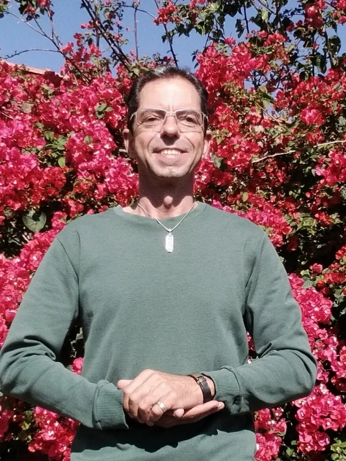 Caio Gracco de pé, com as mãos unidas, diante de uma buganvília em flor